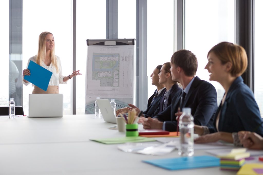 A Woman Stands at the Head of a Table Holding a Presentation Folder While Businesspeople Sit at the Table for a Presentation