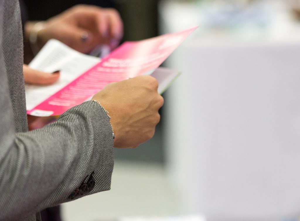 Close Up of a Woman's Torso in a Gray Business Suit Holding a Pink Flyer with White Text on It